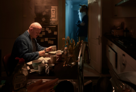 A darkly lit kitchen with on older man sitting at a cluttered table with a masked figure in the doorway.