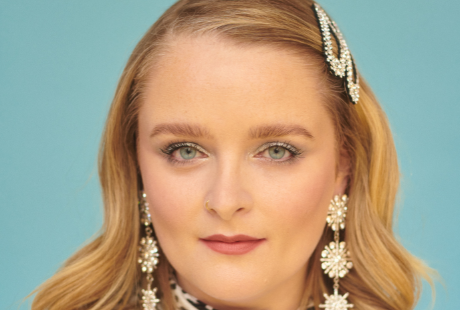 A blonde woman with jewelled earrings and hair clips looks directly to camera in front of a blue background.