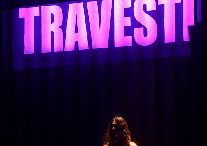 A landscape photograph of dark curtains at the back of a stage with the word "TRAVESTI" lit in large pink letters across the top. At the bottom in the middle, there is a woman visible from the neck up with long hair. Her face is obscured by shadow.