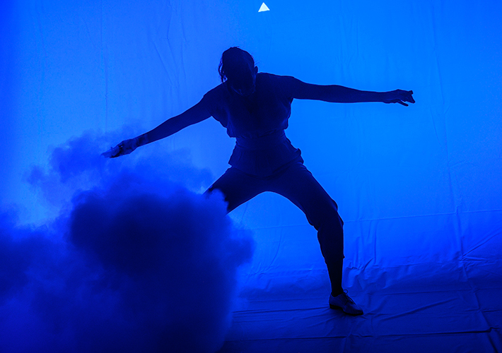 A landscape photograph of a dark blue adult silhouette standing in a blue space. They are facing the camera, though their face is obscured in shadow. Their legs are splayed, with their knees slightly bent, with the leg on the left of the image becoming obscured mid-thigh by a blue cloud rising from the ground. Their arms are out parallel to the floor and they seem to be holding something in each hand. They are slightly hunched. They wear brogue shoes, long socks, three-quarter-length trousers and a tunic with no sleeves which is cinched at the waist.