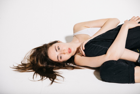The figure of a young woman lying sideways on a white background clutching her knees to her chest.