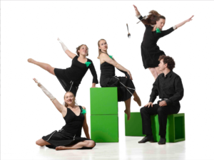 A landscape photo of Darrah Carr Dance, pictured in a still from their piece Melange 445. There are 5 dancers in a white space. Four of the dancers are women who are wearing black tops and skirts, with one male dancer wearing a black long-sleeved shirt, black trousers and black shoes. On the left, one of the female dancers sits on the ground with two green blocks that are slightly taller than her to her side towards the centre of the image. The dancer on the ground is smiling at the camera with her right are raise outward towards the top corner. Behind her, one of the female dancers stands, leaning on the green blocks with her arm and leg raised out. Another sits on the blocks with her legs crossed and her hands on her knees. To the right of this, a silver spoon is falling from the air and another female dancer is jumping up with her arms out. Below her, the male dancer is seated on another green block with a hollow opening.