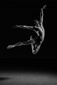 A black and white portrait photograph of Gearóid Solan, an Irish-born dancer, currently dancing with Boston Ballet. He is leaping in the air in a dark room with a grey floor. He is wearing a pair of shorts and his complexion is pale. He is leaping into the air. He is in profile with his face pointed towards the right of the image. One leg is kicked out straight behind him, with his toe pointed. The other leg is bent under him, with his calf and foot pointing up towards his back. He has one arm pointed back, in parallel with his straightened leg. His other arm is up towards the ceiling, with his wrist relaxed and his fingers reaching out.