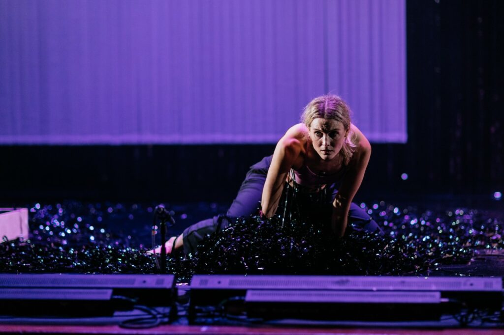 A colored landscape photo of Sibéal Davitt performing an excerpt from her piece Minseach. She is wearing a pink tank top and black fitted pants. She is barefoot and surrounded by piles of black tinsel on a stage with a light purple backdrop. There is a small microphone in front of her along with a row of lights, whose backs face the camera, and she is on her hands and knees holding herself up. Her hair is back and there is a sheen of sweat on her forehead as she stares intently out to the right of the camera, looking afraid with a shadow across the right side of her face. The right side of her body is slightly more forward than the left side of her body like she is crawling towards or away from something. 