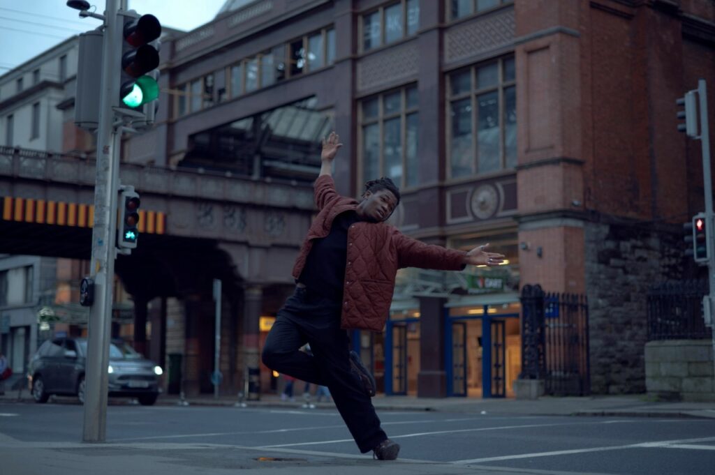 A colored landscape of Tobi Balogun in his piece Ara. Tobi has a deep skin tone with short, dark brown dreadlocks. His entire body is photographed, and he is wearing black pants and a black shirt with a reddish-brown hip-length jacket. His body is facing forward with his head turned to the left. He is next to a traffic light on the edge of the sidewalk. One leg is carrying his weight. The other is bent with his foot behind the other leg. Both arms are extended out with his palms facing forward. His right arm is vertical and his left is horizontal.