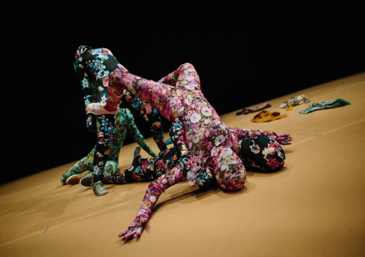 A landscape photograph of four dancers, two male and two females. The dance floor is a beige colour and the background is black. Each of the dancers are dressed in a floral morph suit. One pink, one blue, one black and one green. The floral morph suits are covering each part of their skin from head to toe. In the foreground of the image a female dancer lays on top of one of the male dancers with her face facing upwards with the male dancer lying flat underneath her perched body. Behind them are the other two male and female dancers, one with an arched back and face facing upwards, the other sitting down behind them looking up at their stance. Clothes are scattered on the floor in the background of the image.
