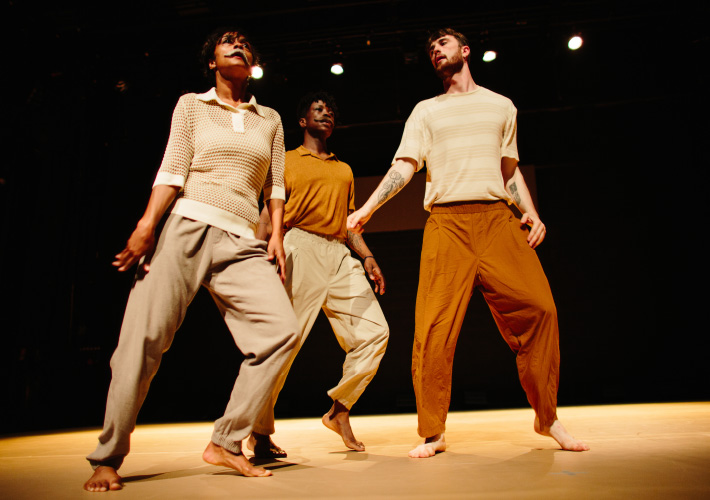 A landscape photograph of three dancers on stage, two males and one female. The dance floor is cream in colour and the background is black. The female dancer is dressed in long cream trousers with a cream mid sleeve polo neck top. The male dancer at the centre of the image is dressed in long cream trousers with a yellow polo shirt. The male dancer on the right is dressed in brown long trousers with a cream and white striped t-shirt. All three dancers are barefoot. The female dancer is wearing a grey moustache.