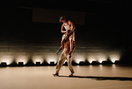 A landscape photograph of two dancers on stage, one male and one female. The dance floor is a cream colour and the background is black. The male dancer is dress in long beige trousers with a brown t-shirt. The female dancer is dressed in a cream top with cream shorts. The two dancers are caught mid-flight, walking across the stage. The female dancer is on the male dancer’s shoulders, holding his face with both hands to secure herself. The male dancer’s arms are by his side.