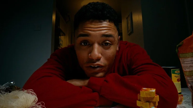 A landscape colour photograph of Irish-Sudanese artist Adam Mohamed. He is pictured in the centre of the image from head to shoulders with his arms crossed on a table in the foreground. His brow is furrowed and his mouth is slightly open. He wears a long-sleeved red jumper. Behind his shoulders is a hallway with a picture frame on one wall. On the table around him are culinary items like dry noodles and stock cubes.