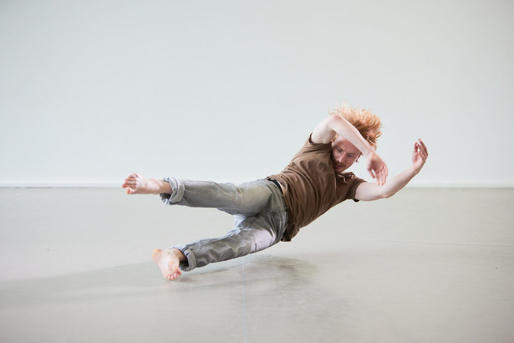 In this photo, a man in a brown short sleeve tshirt, blue jeans and bare foot is horizontal to the floor elevated inches from the floor. He is looking down towards his feet and his arms are bent near his forehead. His hair is extending from his head as if he was falling.