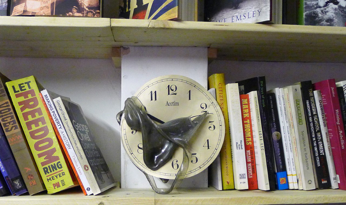 A set of shelves holds a number of anarchist/left books and magazines that have a light coating of dust/ash. At the centre of the image is a clock on which the clear plastic cover has melted, making a soft-edged, grey form on the face of the clock.