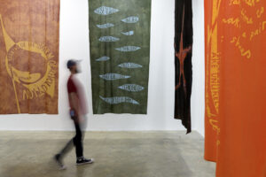 A blurry figure walks through the gallery in front of large banners suspended from the ceiling. They are brown, green, black and orange in colour. The green banner is the only one that is fully visible. Text on it, made through the process of batik, with its characteristic crackle pattern, is written inside of pale blue shapes that resemble wounds, slits, or leaves. The text reads: Those you see lying here, buried in mud As if they lay already in their grave – They’re merely sleeping, are not really dead Yet, not asleep, would still not be awake.