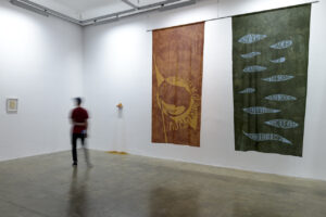 A blurry figure walks through the gallery in front several works: a light coloured work framed in white, a red book, acting as a shelf, covered in brown powder that spills onto the floor, and two large banners suspended from the ceiling. The banners have the characteristic crackle pattern of batik. One is brown with a gold pattern and reads "We already hear the roaring of the storm of world revolution." The other is a muddy green with blue shapes. It reads "Those you see lying here, buried in mud; As if they lay already in their grave –; They’re merely sleeping, are not really dead; Yet, not asleep, would still not be awake."