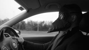 Landscape black and white close up photograph of a man wearing a raven plague mask driving a car holding the wheel.