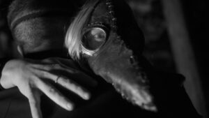 Landscape black and white close up photograph of a woman with a blonde bob in a raven plague mask looking forwards whilst hugging a man with his back turned to the camera.