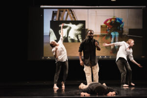 There are 4 performers on stage: one white female, two Black male performers,one white male. 2 of the performers are wearing black button-down shirts rolled at the sleeves with tan pants and the two white performers are wearing white button down shirts rolled at the sleeves with black trouser. 3 of the performers are standing and one is laying face down on the floor. Behind them is a video screen showing their movement in one box overlaid with the image of a white woman sitting in a chair. Her image is distorted.