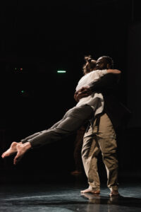 There are two dancers embracing each other on stage - one white female and one Black male. The Black dancer is swinging the white dancer around and the white dancer’s legs have lifted off the floor and extended. She is face down on his shoulder.