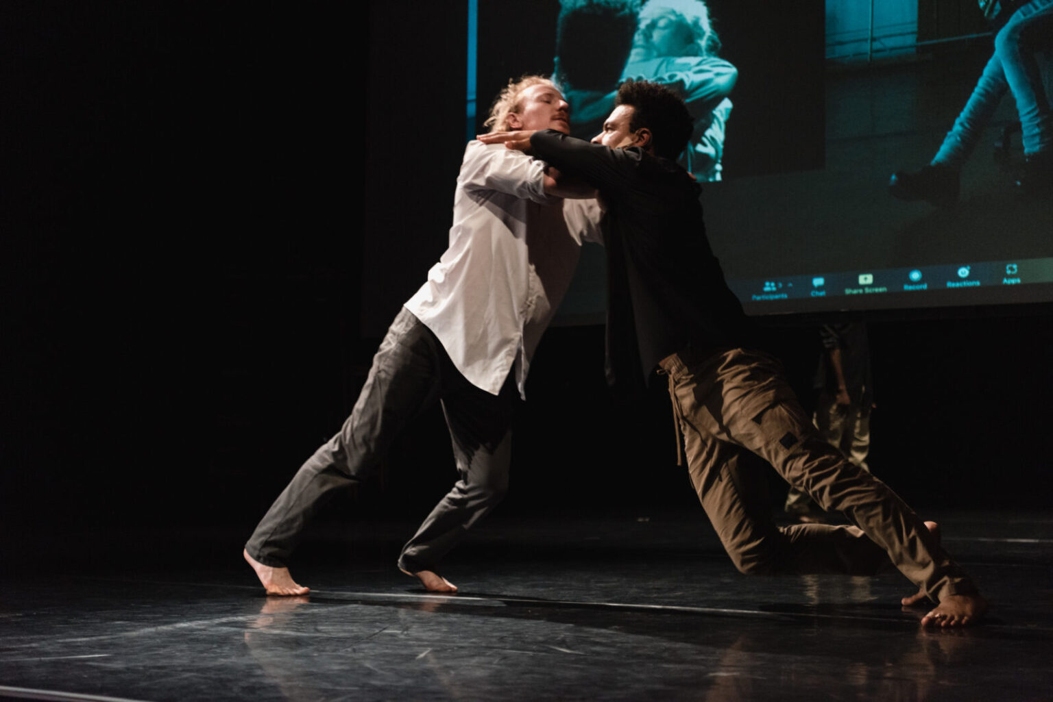 There are two dancers on stage: one white male in a white button-down shirt rolled up at the sleeves and black trousers and one Black male dancer in a black button-down shirt and tan pants with the sleeves rolled to the elbow. Both dancers are barefoot They are leaning into each other facing each other with their hands around each other’s necks with support on bent elbows.