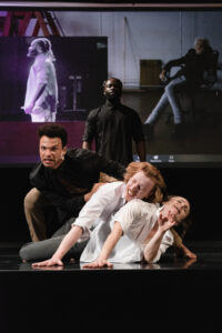 There are four dancers on stage standing in a line. The front two dancers male and female are both white. The back two male dancers are Black. The first dancer in the line is lying on the floor with her head lifted facing the audience with an expression of pain. The second dancer in the line is leaning over her with his head nearly resting on her shoulder and facing the audience. He is smiling with his eyes closed. The third dancer in line has two hands on the back of the second dancer. He is leaning over the to the right. The 4th dancer is standing fully upright back at a distance from the first three dancers. There is a video screen behind the group. One box on the video screen shows the 4th dancer in the line but his at his profile and to the left of the screen is an older white woman sitting in a chair