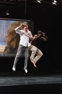 On stage, there is one white male dancer jumping in the air with his legs extended beneath of him and a second Black male dancer who is trying to bump into the first male dancer. The second dancer is also in the air. His knees are pulled up towards his chest. Behind both dancers is a video screen. On the screen is a picture of an older white woman looking out a window.