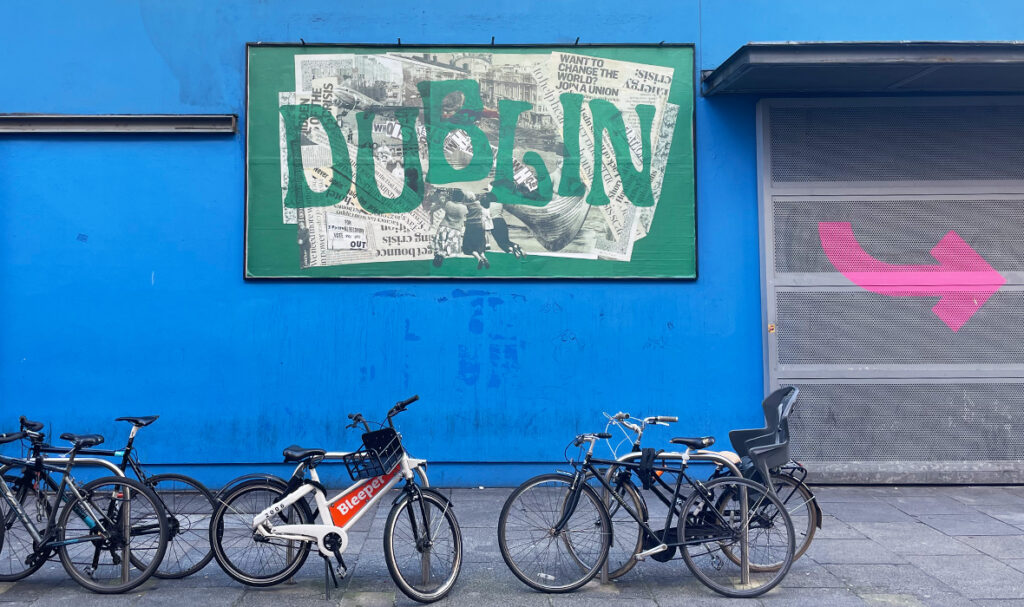 A photo of the billboard in front of Project Arts Centre. The billboard features an emerald green frame with paper collage in the centre. The collage shows newspaper clippings of headlines oncerning inflation, the rising cost of living, the housing crisis, and other public sphere isses concerning residents in Dublin frame black-and-white imagery of Dublin City, with historical and political figures is layered over one another. In the centre in large type is “DUBLIN” in transparent green lettering.