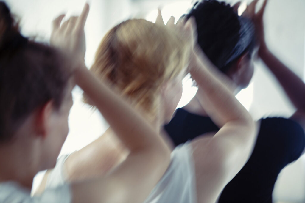 Landscape colour, motion blurred close up photograph of three women’s head and shoulders standing in a diagonal line with their right arm raised and rested on each other’s head, photographed from behind, taken at the Grand Studio, Brussels.