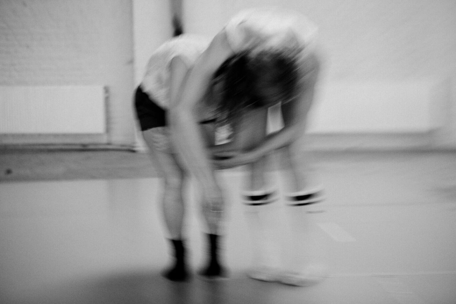 Photo2_GrandStudioResidency – Justine Doswell Landscape black and white photograph of two standing dancers in parallel position, standing next to each other centre frame with their backs curved over towards the floor in an embrace, against a white background taken at the Grand Studio, Brussels.