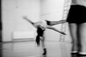 Landscape black and white, motion blurred photograph of a dancer wearing black shorts and a white t-shirt in a layout position, left leg extended, torso horizontal to the floor accompanied by another dancer standing stage right in foreground, their torso cropped at waist with a ladder in the background, taken against a white background at the Grand Studio, Brussels.