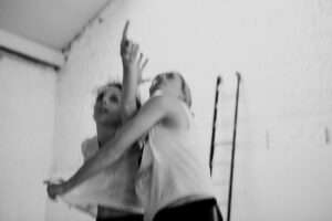Landscape black and white, motion blurred photograph of two dancers wearing black shorts and white t-shirts, cropped at their hips. Dancer on stage right pointing upwards on a diagonal with their left arm downstage left, whilst dancer on stage left embraces with their right arm and pulls t-shirt with their left arm, with a ladder in the background against a white background taken at the Grand Studio, Brussels.