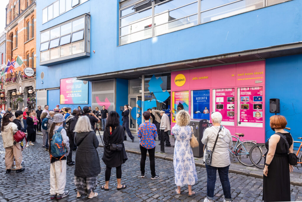  The landscape image shows a large audience gathered outside the Project watching Tobi Balogun and dancers perform. The top of the image shows the blue exterior of the building and the pink poster boxes. In the bottom half of the image, we see the back of the audience. They are standing on the cobbled street. 