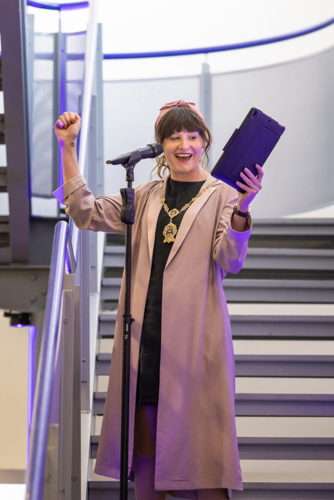 The portrait image shows deputy Lord Mayor Darcy Lonergan. She is standing on the staircase with a microphone stand in front of her. She has brown hair tied up with a pink headband on. She is wearing a black dress and a long beige coat. She is also wearing the Lord Mayor’s chain. She is looking towards her left hand which holds an ipad. She is raising her right fist in the air and is smiling. 