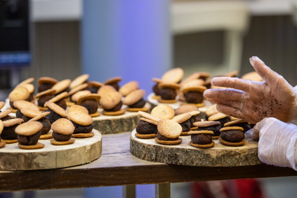 The landscape image shows three wooden platters, made out of tree trunks, sitting on a table. Each one holds approximately 10 little cakes. Each cake has a beige coloured biscuit underneath and on top, of a darker chocolate coloured centre. There are two hands to the right of the image arranging the cakes. The hands are wearing white plastic gloves. 