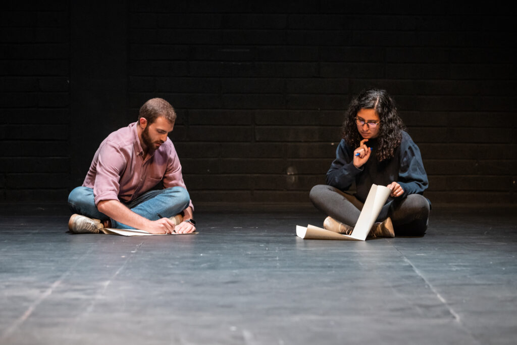 Two people sit on the floor with their legs crossed. They are writing on large sheets of paper. The background and floor are black. They are in a Theatre space. A man with short brown hair and a beard is sitting on the left. He is wearing a light pink shirt, blue jeans and white trainers. A woman with long black curly hair sits on the right. She is wearing a navy sweatshirt and tight black jeans. She is holding a pen and her right hand is resting under her chin.