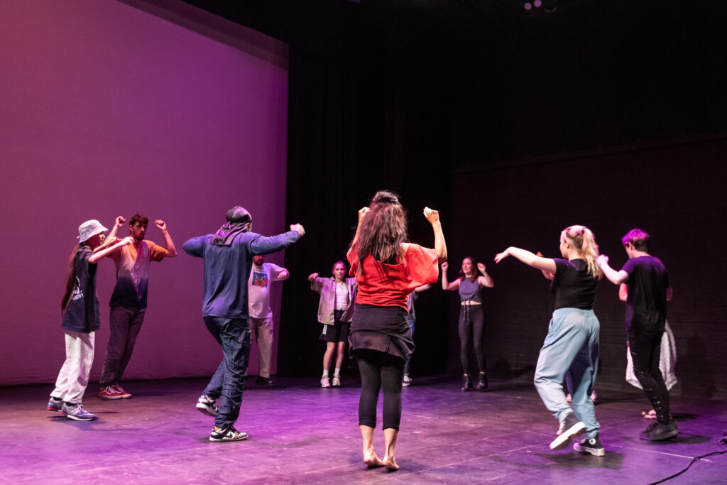 12 people are dancing in a circle. They are facing towards the centre. They all have their hands in the air at shoulder height. Their hands are in fists. Some of them are balancing on one leg. In the foreground we can see artist Tobi Omoteso. He is dressed in black. To his right there is a woman with black leggings, a black skirt and a red t-shirt. She has long black curly hair. To his left there is a child with long black hair, a black t-shirt, grey tracksuit bottoms and a grey bucket hat. The background of the image is black and dark purple. They are in a theatre space. 