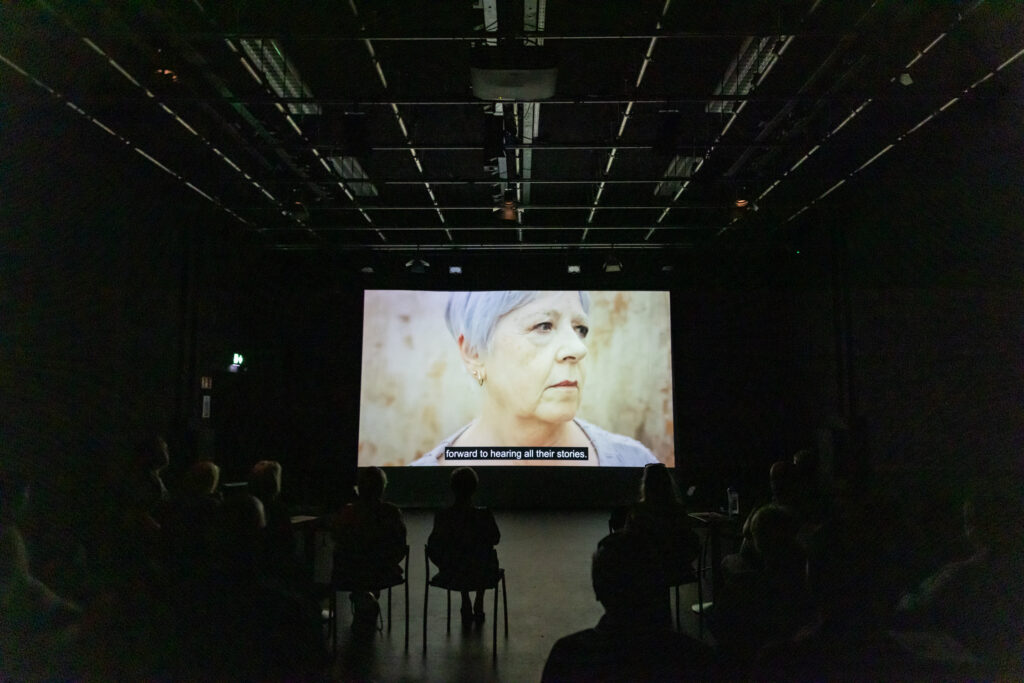 The landscape image shows a seated audience watching a film. The audience is sitting in the dark. In the centre of the image you can see the film screen. On the screen is a close up of a woman's face. She has a serious expression and is looking to her left. She has light purple hair and a purple top. Above the screen we can just about see the lighting rig. Below the screen there are silhouettes of chairs and audience members with their backs to us.