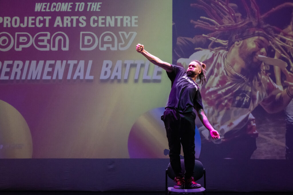 This landscape image shows dancer Dylan Chapman performing in the Space Upstairs. He is standing on a chair in front of a screen. On the screen behind him is an image of a dancer and text that reads ‘Welcome to Project Arts Centre Open Day, Experimental Battle’. Dylan is wearing black jeans and a black t-shirt. He has dreadlocks that are tied up on top of his head. His facial expression is aggressive. He has his right hand in a fist and his right arm extended. His left arm hangs down by his waist. 