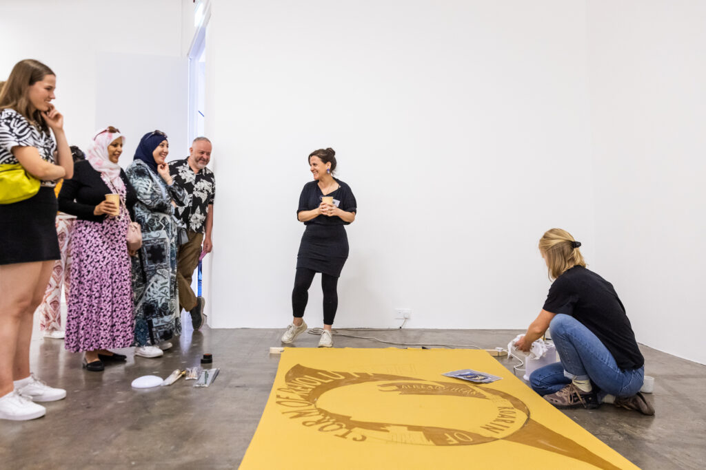 The landscape image shows a demonstration in the gallery. Visual Arts curator Sara Greavu stands in the centre of the image. She is wearing black leggings, a black skirt and a black top. She has black hair tied up in a bun. She is smiling and looking to her right. On the floor there is a large piece of yellow material. Artist Mary Kervick is kneeling down beside it, demonstrating Batik. She is wearing blue jeans and a black top. She has blond shoulder length hair. To the left of the image there are four people observing the artist. They are all smiling. 