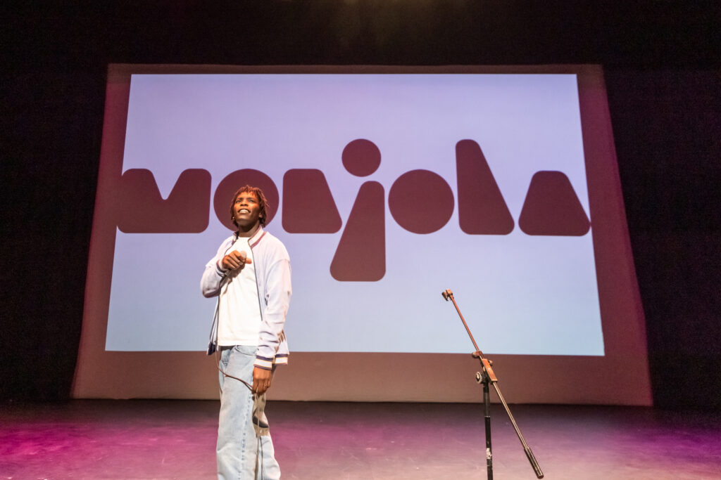 The landscape image shows music artist Monjola performing in the Space Upstairs. Behind him is a projected backdrop with his name spelled out in a large scale black font. He is a black man with short black hair. He is wearing blue jeans, a white t-shirt and a light blue bomber jacket. There is a microphone stand in the foreground. He is holding a microphone in his right hand and is smiling up at the audience.  