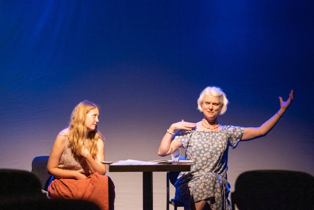 The landscape image shows Róisín Power Hackett and Emilie Conway giving a talk in the Cube. Róisín is sitting on the left and Emilie is on the right. Róisín  is a white woman with messy long blonde hair. She is wearing a coral pink long skirt and a short sleeved patterned t-shirt. Emilie is a woman with a pale complexion and a loose blonde bob. She is wearing a blue dress with flowers. She has her right hand placed on her right shoulder and her left arm in the air with her palm facing the sky. Between them there is a square wooden table with papers and a glass. 