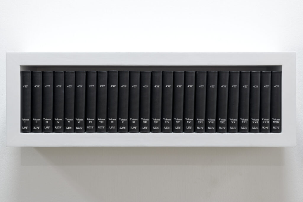 A white shelf holds 24 black hardback books with silver printing on their spines. Each reads 4’33” and the volumes are numbered 1-24 in Roman numerals. Each has 11.375” printed at the bottom of the spine.