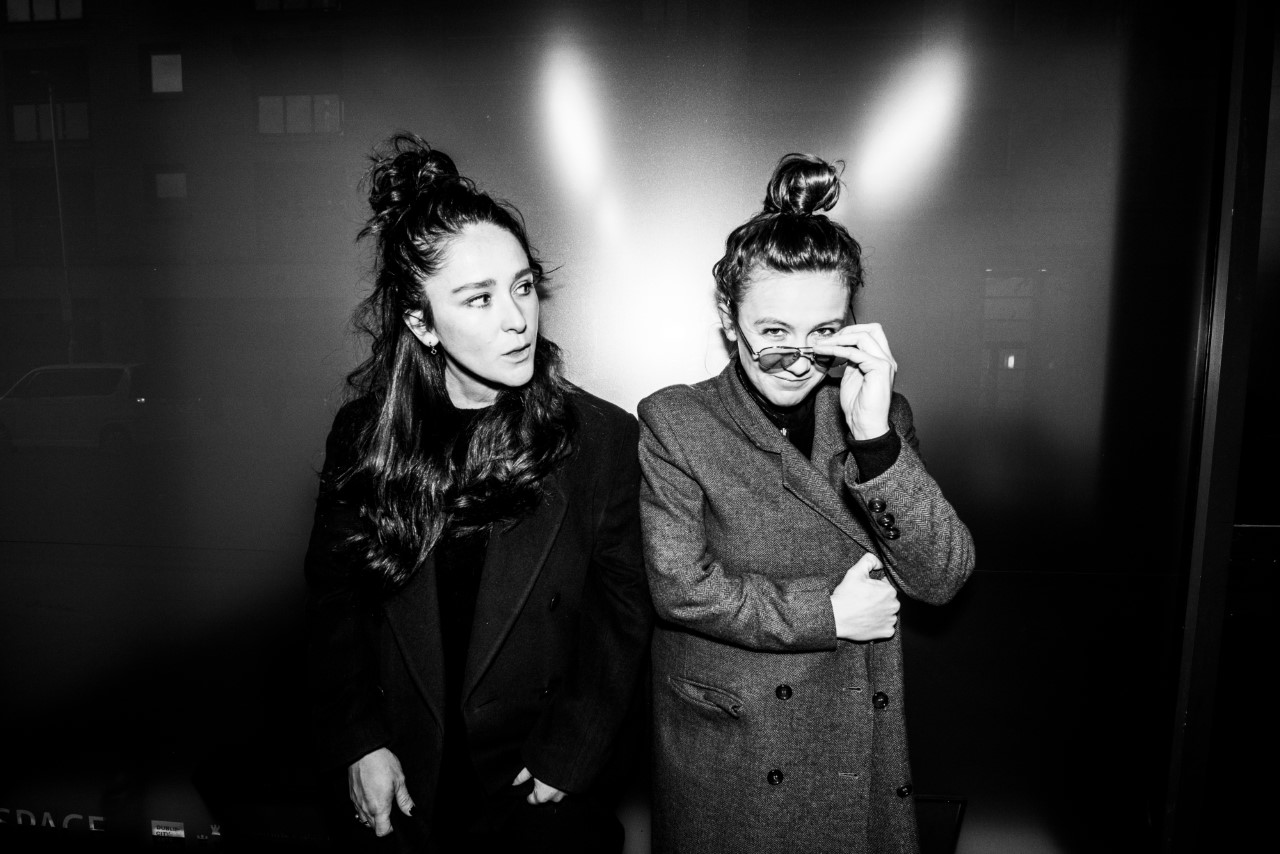 Black and White image of two women, Maud (on left) and Zoe (on right) Maud is looking to the right and leaning forward slightly Zoe has glasses on and she is lowering them down her nose and smiling while looking at the camera. They are standing against a plain background Both are wearing outdoor coats