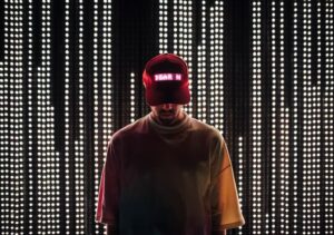 A white man wearing a tie-dye shirt and red cap that says ‘Dear In’ on it in pink LED lights. He is standing in front of a wall of strings of white LED lights.