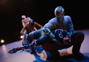 A colour landscape photo of 4 dancers on an empty stage with a black background. All the dancers wear full-body floral morph suits with clothes over them. In the foreground, one dancer is crouching with a second dancer's head suspended across his lap. The second dancer has their feet planted on the floor, knees bent, with their chest and face too the ceiling and their left arm suspended out to the side. In the background, one dancer is bent over with their hands on the second dancers knees. Behind them, one dancer is standing up and leaning backwards behind them. The four dancers stretch back in a line across the image.