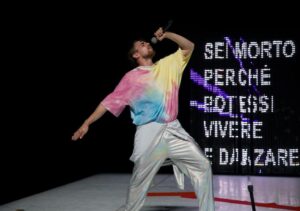 A white man wearing metallic silver pants with a tie-dye shirt standing in front of LED lights that spell ‘Sei Morto Perche Potessi Vivere E Dinvare’