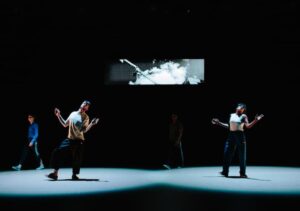 A colour photo of four dancers on a stage wearing tracksuit bottoms and plain t-shirts of different styles. The stage is lit in two overlapping points, with the rest of the floor and wall in darkness. On the back wall, there is a landscape image being projected showing a crane and a cloudy sky in black and white. In the two points of light on stage, two dancers stand separately. They face the camera, with their left feet planted on the stage and their right heels on the stage in a slight step out, toes pointing up. They have their arms hang down from their shoulders but are bent out and upwards at the elbows, with their hands splayed out and facing the camera. Their faces are angled slightly upwards towards the ceiling. In the dark behind them, two dancers walk separately across the stage in the same direction.