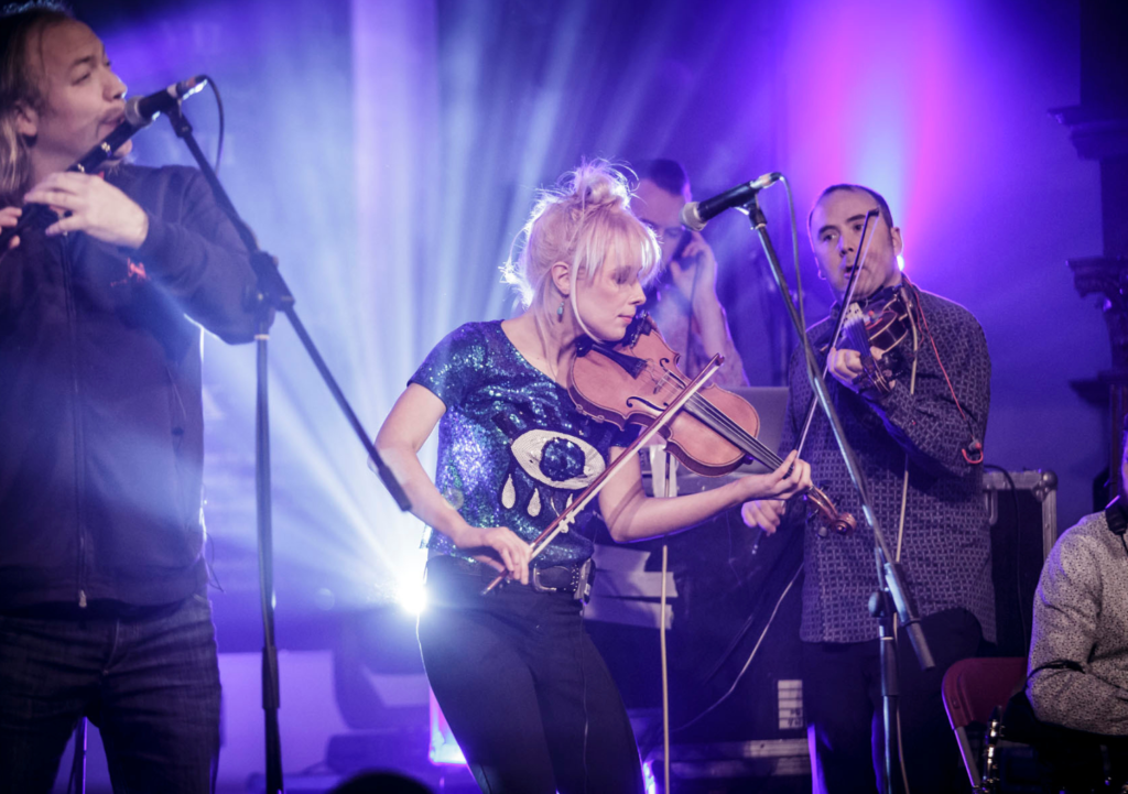 A band is on a stage. On the left there is a man playing a flute into a microphone.He is wearing a black hoodie. In the middle there is a woman playing a fiddle. She has her blonder hair tied up and is wearing a sparkly top. On the right there is a man playing a fiddle. He is wearing a shirt. In the very background there is a glimpse of a DJ standing at a set of DJ decks wearing headphones.