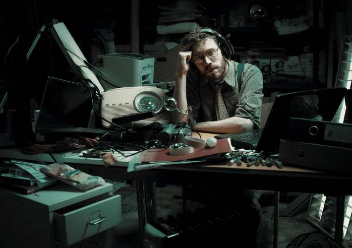 A colour photo of a white man with brown hair wearing glasses. He is looking deadpan into the camera. He is positioned in the centre of the image, sitting at a desk in a room in which he is surrounded by computers and phones from the 90s.