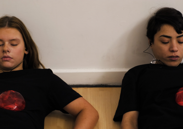 Two young white women together closely; we see their bodies from the shoulders up. One has a short black pixie cut and the other has a brunette bob cut. They are both sitting with their eyes close wearing black shirts with one red heart each over their chests.