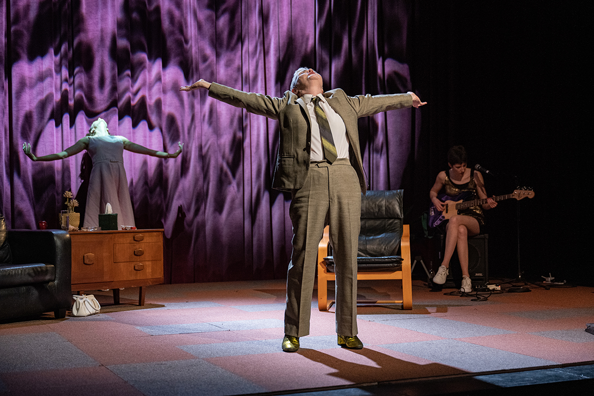 A white woman with short hair wearing a green-grey suit and green tie stands with her arms outstretched and face lifted upwards. Behind her, there is a couch, wooden sideboard and black leather chair. A white woman in a pink dress mimics her stance against a red curtain which has projections of smoke. To the right of the image, a white woman with short, dark hair sits on a guitar amp playing a blue bass guitar.