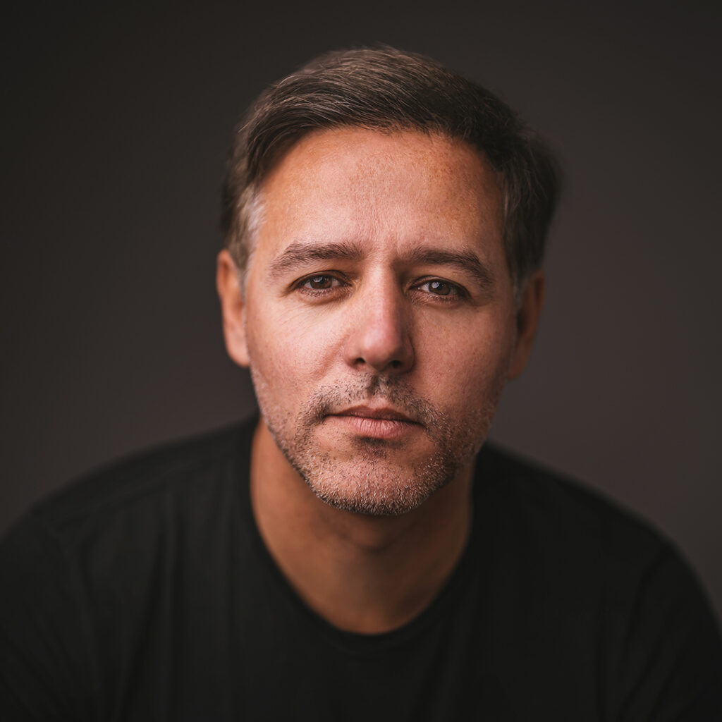 A colour portrait photo of Alejandro Llosa. He is pictured from the shoulders up, leaning towards the camera in a dark room, with his face in focus. He has short hair and light stubble. He looks into the camera with a slight smile. He is wearing a black t-shirt.